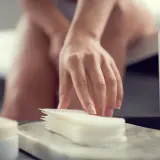 Dłoń kobiety sięga po wkładki higieniczne. W tle widać fragment ciała i butelkę z płynem. Może to być związane z brązową wydzieliną z pochwy.