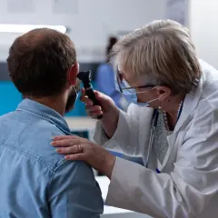 Lekarka w masce bada ucho pacjenta otoskopem. To wizyta u specjalisty z zakresu otorynolaryngologii.