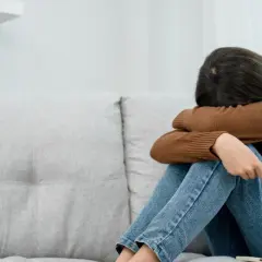 Kobieta siedzi skulona na kanapie, przytulając kolana. Jej twarz jest ukryta, co może symbolizować depresję lękową i poczucie osamotnienia.