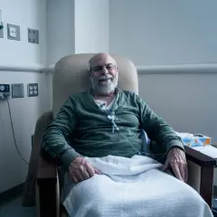 Uśmiechnięty mężczyzna w fotelu szpitalnym, podłączony do kroplówki, otrzymuje żywienie pozajelitowe.