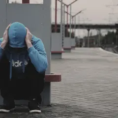 Chłopak w niebieskiej bluzie z kapturem, z rękami na głowie, siedzi na peronie. Może to być efekt działania lsd, wywołujący dezorientację.