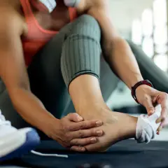 Kobieta masuje skręconą kostkę, obok leżą buty sportowe.
