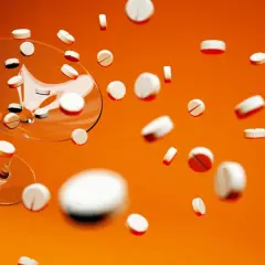 Białe tabletki, w tym Xarelto 20 mg, spadają z kieliszka na pomarańczowym tle.