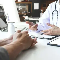 Lekarz omawia z pacjentem leczenie anty HCV. Na stole leżą tablet, laptop i biała buteleczka.