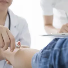 Lekarz aplikuje maść detreomycyna na skórę pacjenta. W tle inny lekarz notuje coś w zeszycie.