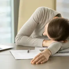 Kobieta z głową opartą na ramieniu, przy biurku z laptopem i dokumentami, w stanie biurowego marazmu.
