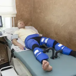 Mężczyzna leży na łóżku rehabilitacyjnym z założonymi rękawami do drenażu limfatycznego na nogach.