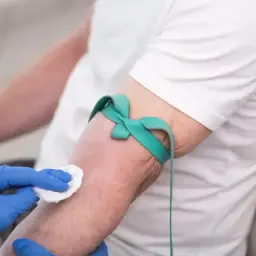 Ręka pacjenta z opaską uciskową i wacikiem, przygotowywana do pobrania krwi. Badanie hbs.