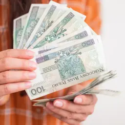 Dłonie trzymające stos banknotów 100 zł. Może to być symbol oszczędności, wypłaty lub nawet hipotetyczna "renta alkoholowa".