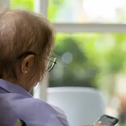 Osoba po chemioterapii w okularach siedzi przy oknie, trzymając telefon.