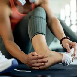 Kobieta masuje skręconą kostkę, obok leżą buty sportowe.