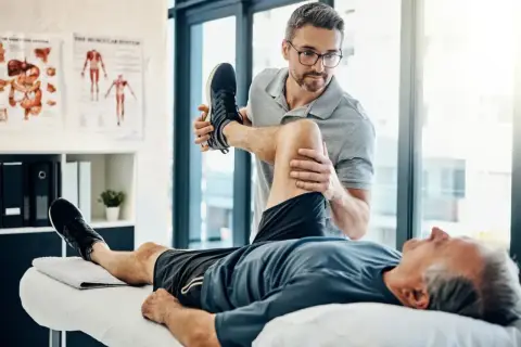 Fizjoterapeuta pomaga starszemu mężczyźnie w ćwiczeniach. Dobre ubezpieczenie zdrowotne zapewnia dostęp do takiej rehabilitacji.
