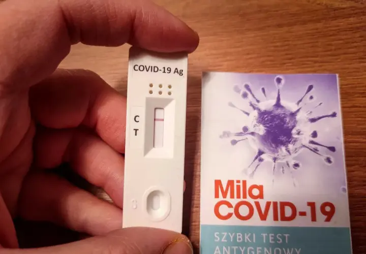 Ręka trzyma test antygenowy COVID-19 z jedną kreską. Obok opakowanie z grafiką wirusa i napisem "Szybki test antygenowy".