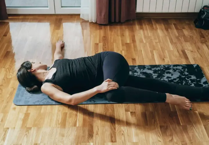 Kobieta w czarnym stroju ćwiczy na macie, wykonując skręt tułowia leżąc na plecach.