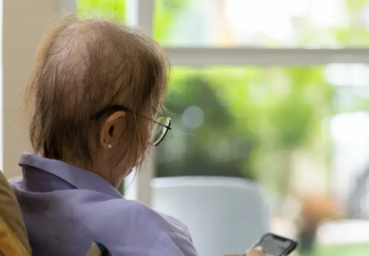 Osoba po chemioterapii w okularach siedzi przy oknie, trzymając telefon.