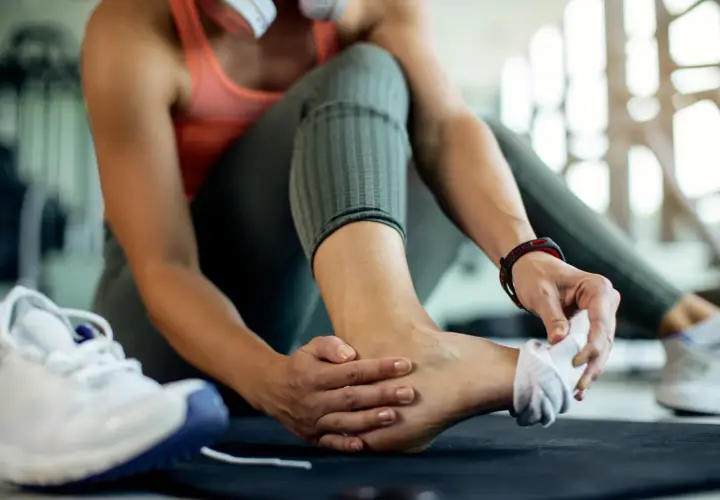 Kobieta masuje skręconą kostkę, obok leżą buty sportowe.