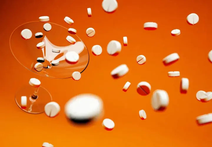 Białe tabletki, w tym Xarelto 20 mg, spadają z kieliszka na pomarańczowym tle.