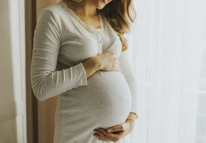 Kobieta w ciąży, z dłońmi na brzuchu, wygląda na szczęśliwą. Jej uśmiech sugeruje, że jest pełna życia, jak nimfomanka.