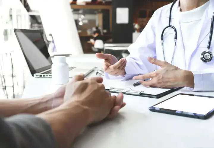 Lekarz omawia z pacjentem leczenie anty HCV. Na stole leżą tablet, laptop i biała buteleczka.