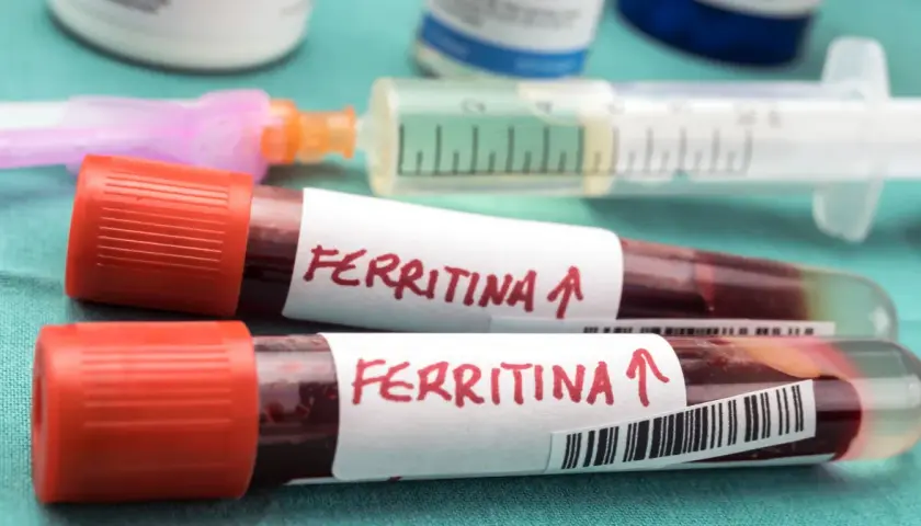 Dwie probówki z krwią oznaczone "Ferritina" i strzałką w górę, sugerujące potrzebę podniesienia ferrytyny. W tle strzykawka i fiolki.