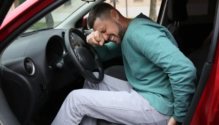Mężczyzna w samochodzie trzyma się za pośladek i nogę, wyrażając ból.