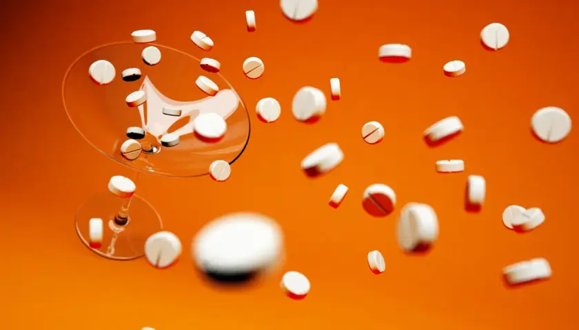 Białe tabletki, w tym Xarelto 20 mg, spadają z kieliszka na pomarańczowym tle.