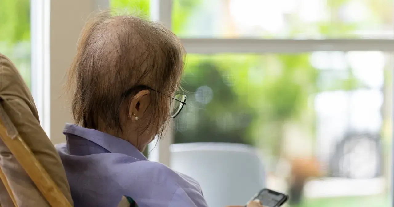 Osoba po chemioterapii w okularach siedzi przy oknie, trzymając telefon.