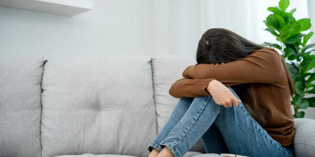 Kobieta siedzi skulona na kanapie, przytulając kolana. Jej twarz jest ukryta, co może symbolizować depresję lękową i poczucie osamotnienia.