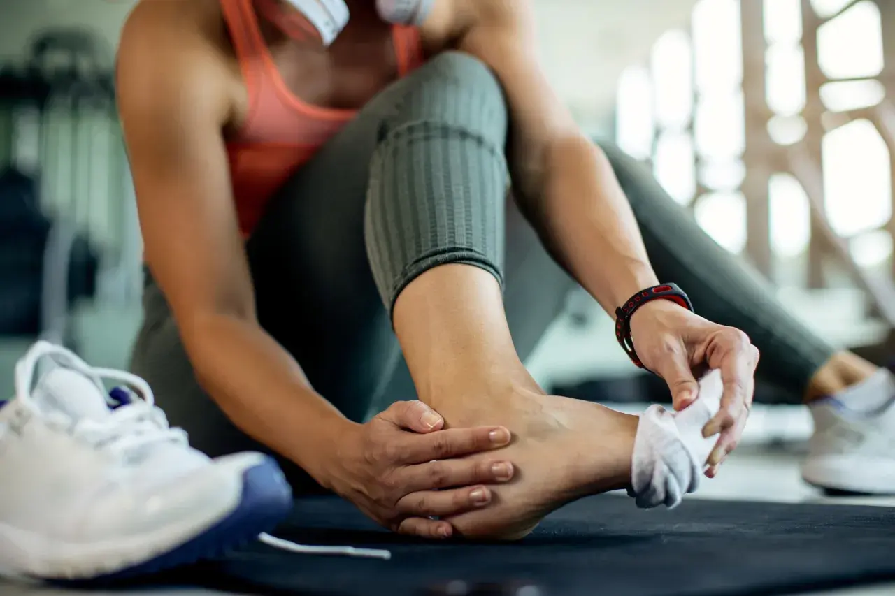 Kobieta masuje skręconą kostkę, obok leżą buty sportowe.